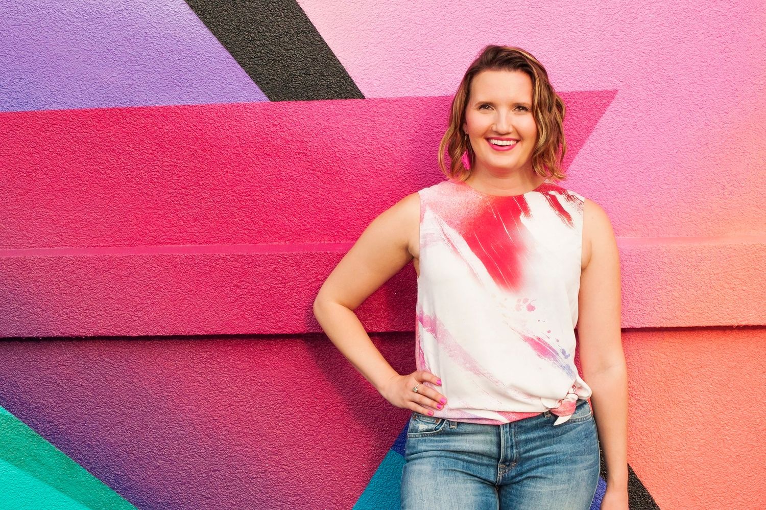 Smiling woman in a white and pink sleeveless top posing against a colorful pink and teal geometric wall.