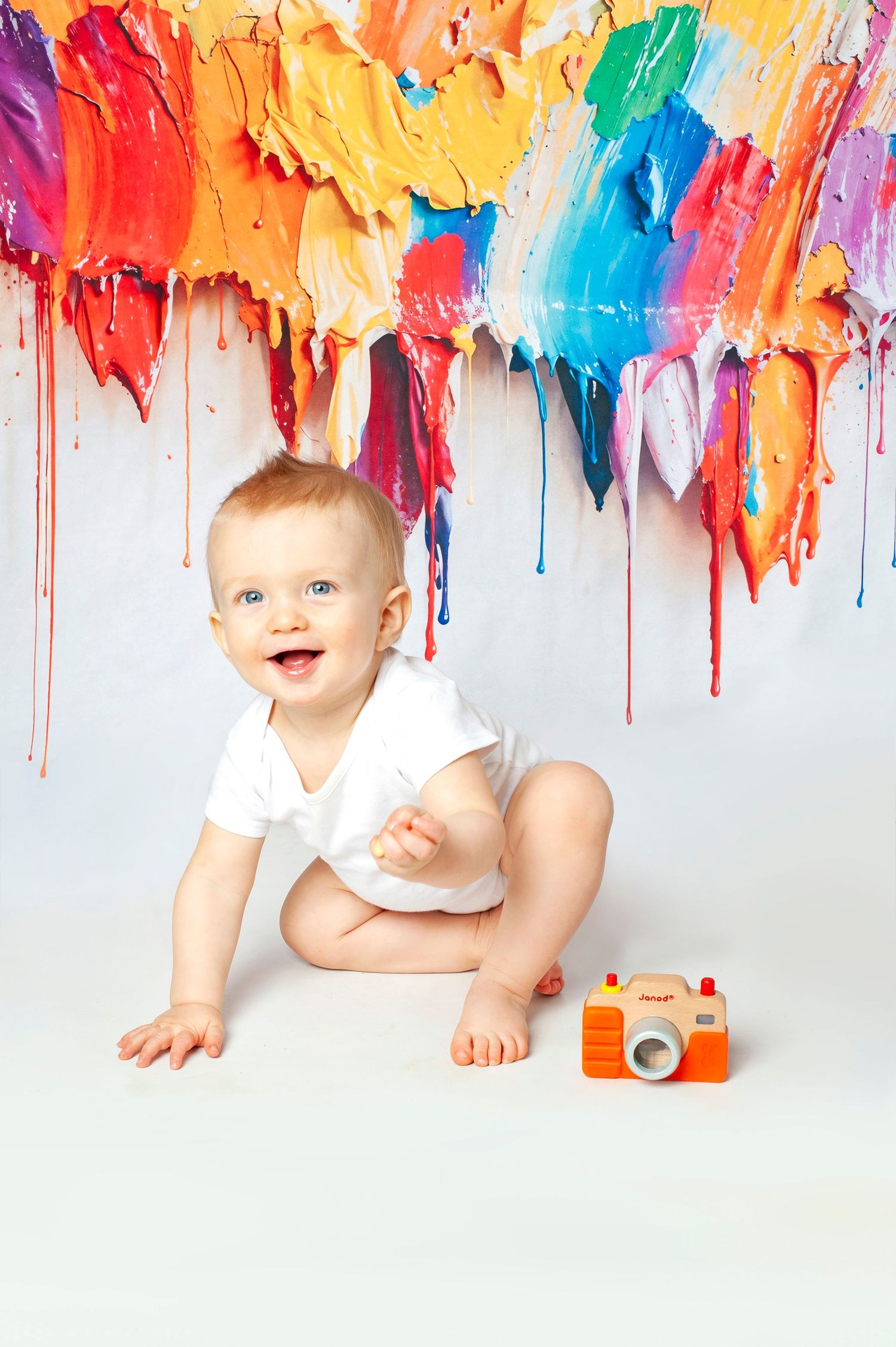 Happy baby in white onesie crawls near toy camera with colorful paint strokes dripping on white background.