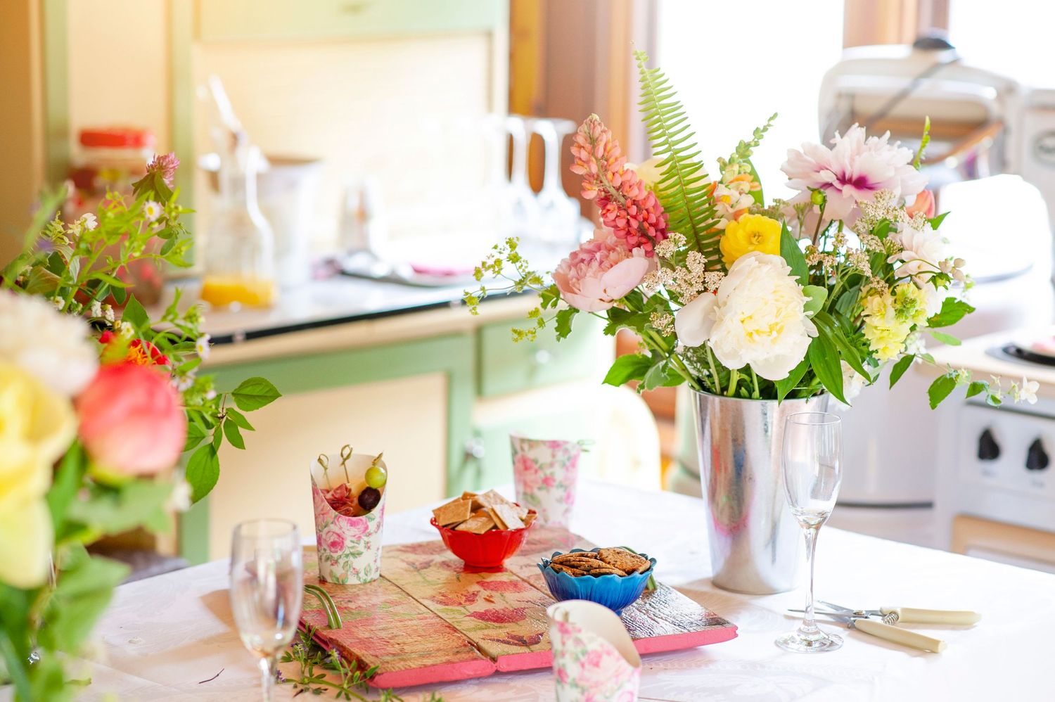 Bright mixed flower bouquet in silver vase on a decorated table with snacks and glassware in a cozy kitchen.