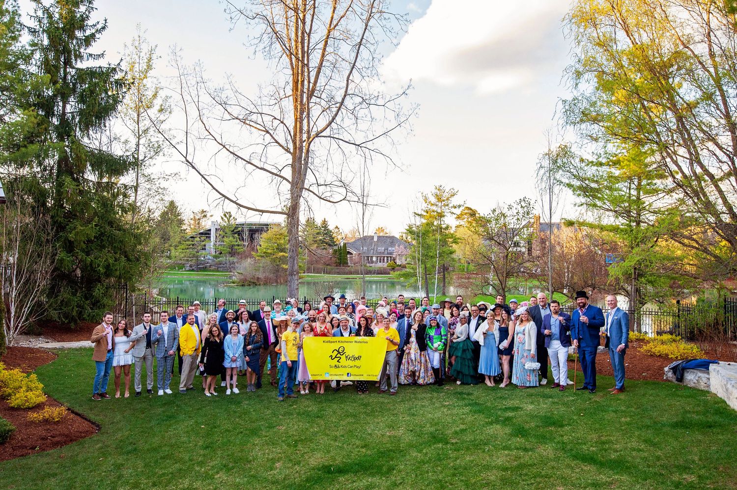 Large group of people posing outdoors on green lawn holding yellow banner near trees and landscaped garden.