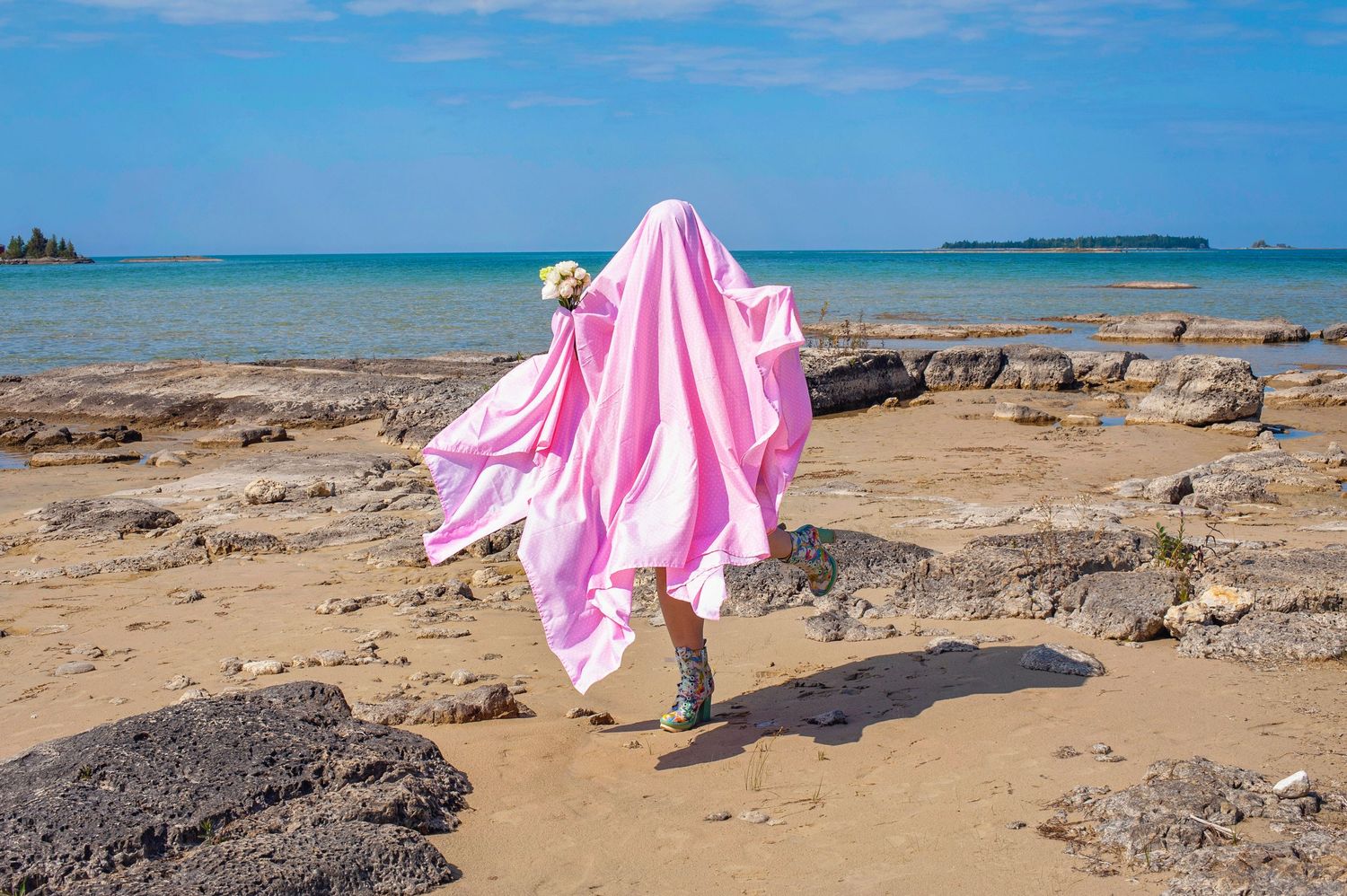 Person wrapped in flowing pink fabric on a rocky beach with turquoise ocean and blue sky backdrop.