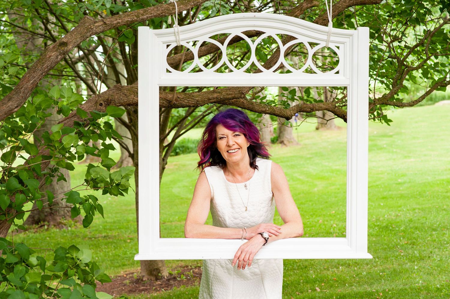 Smiling woman with purple hair posing through decorative white frame in garden.