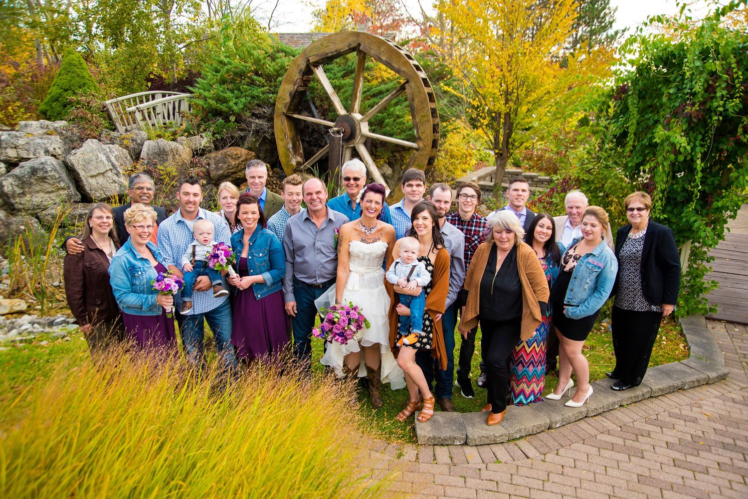 Large wedding party group photo taken outdoors near a decorative water wheel in fall.