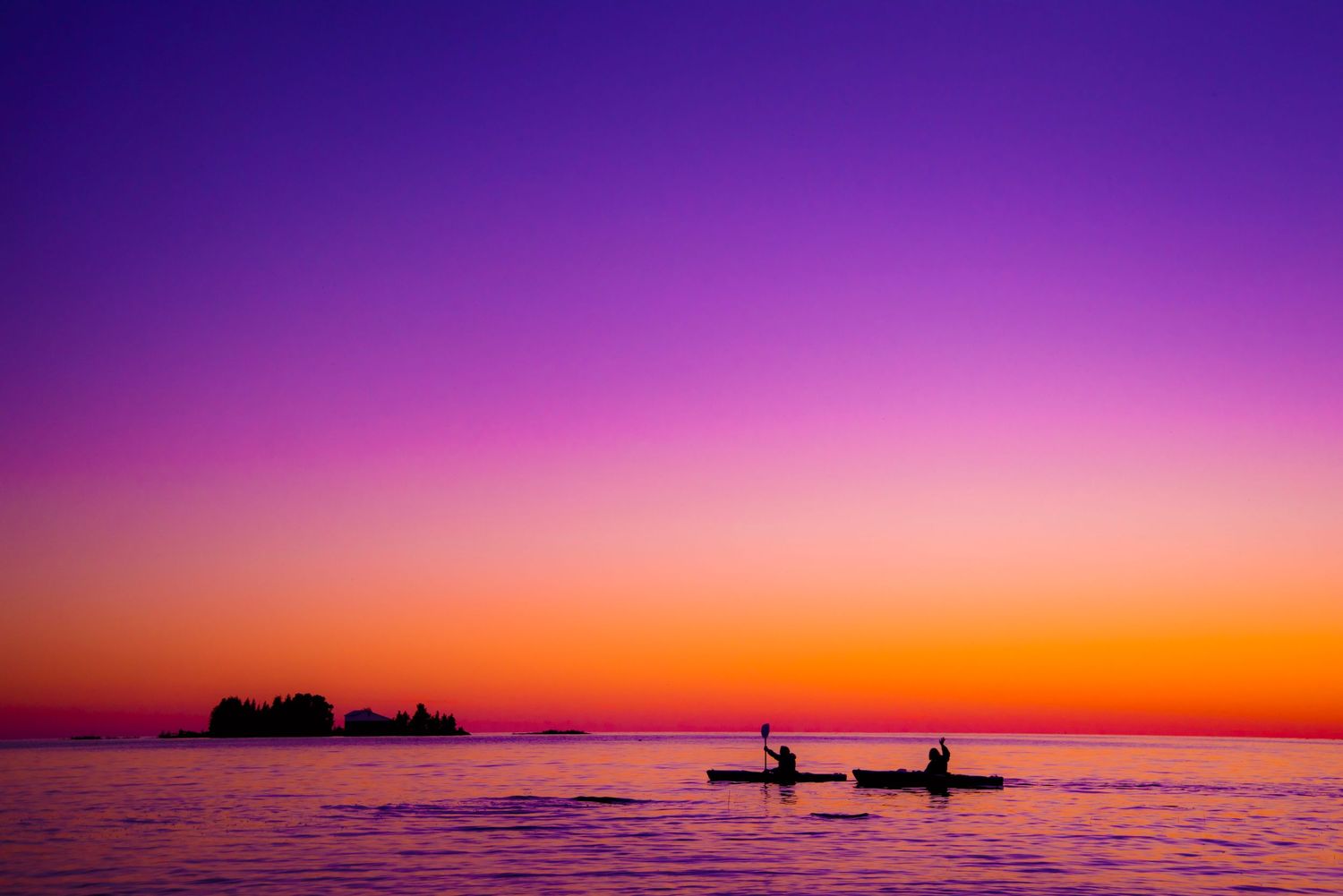 Silhouettes of two kayakers paddling on calm water beneath a stunning purple and orange sunset sky.
