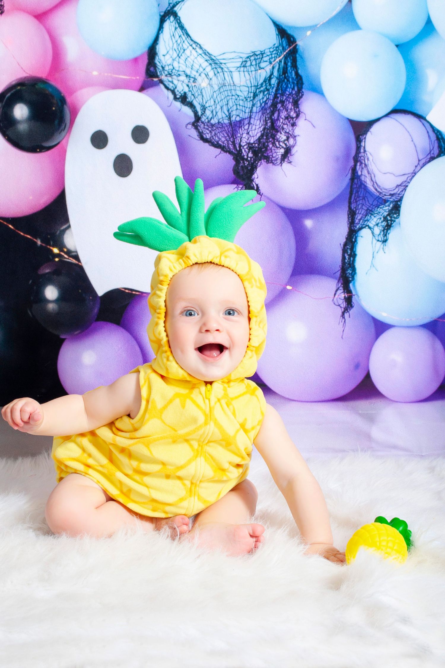 Baby in yellow pineapple Halloween costume sitting on white fur rug with colorful balloons and ghost decoration.