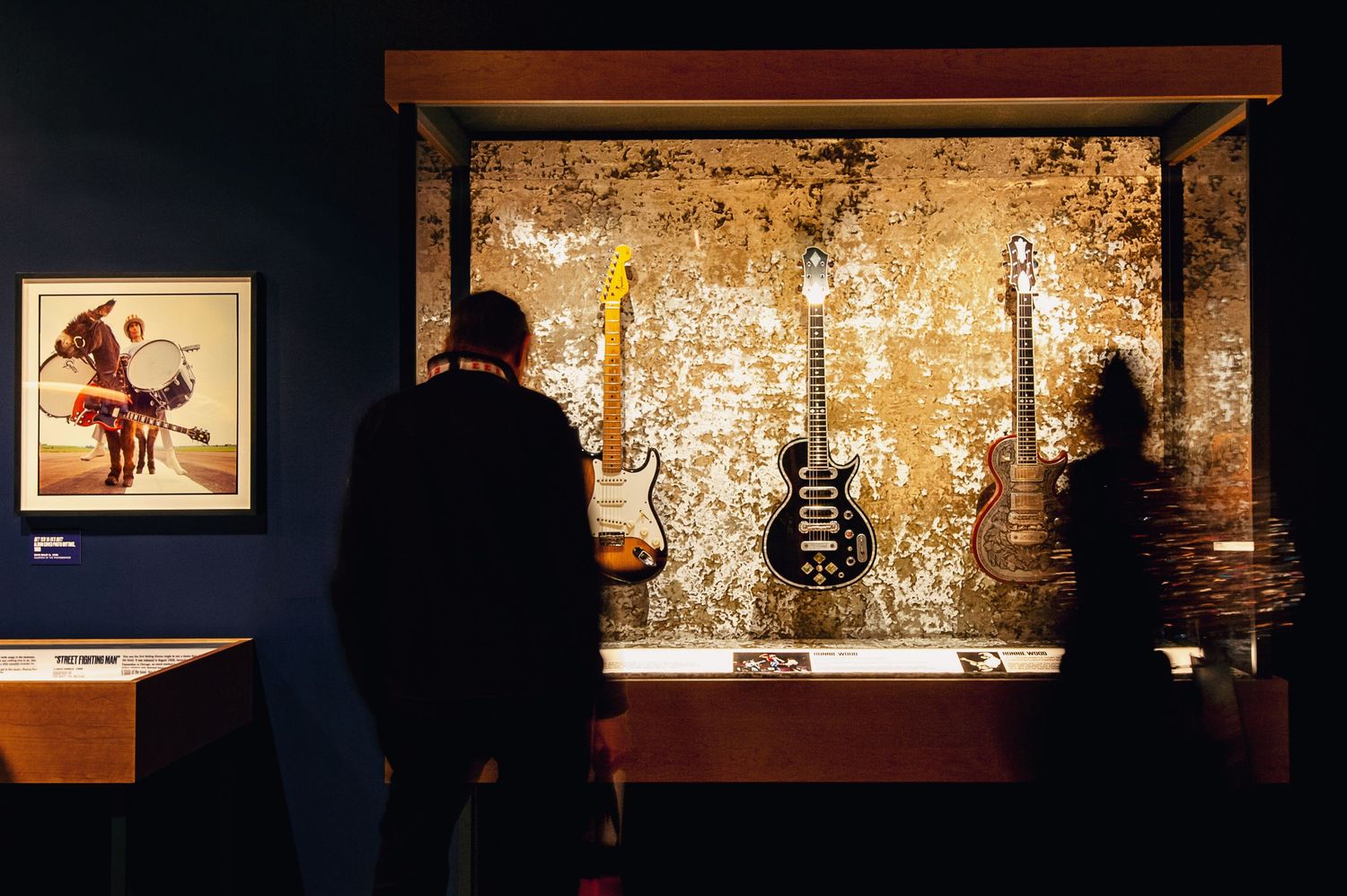 Museum visitors examine vintage electric guitars displayed in a glass case.