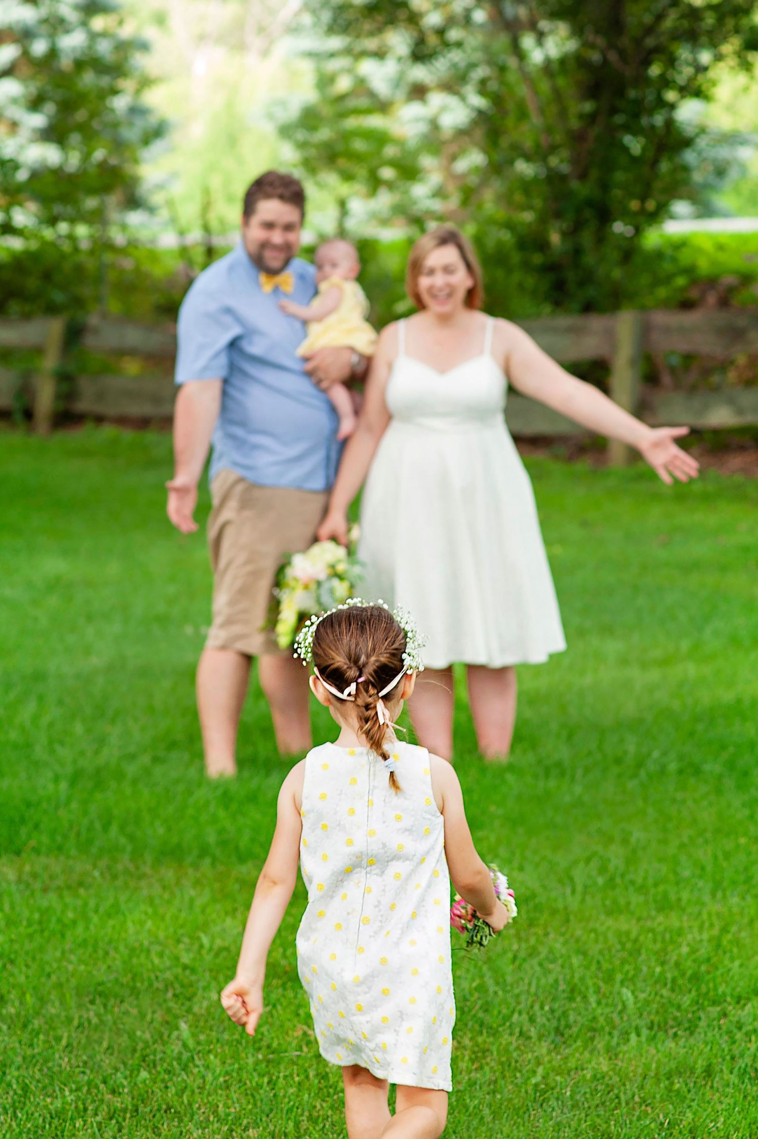 Little girl in white dress with flower crown runs toward her welcoming parents on a sunny green lawn.