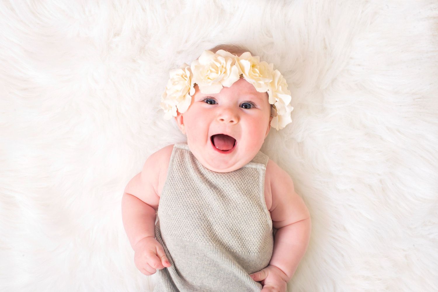 Happy smiling baby wearing a floral headband lying on a fluffy white fur rug.