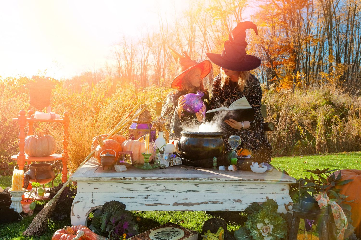 Two women dressed as witches brew potions outdoors in golden autumn sunlight with Halloween decorations.