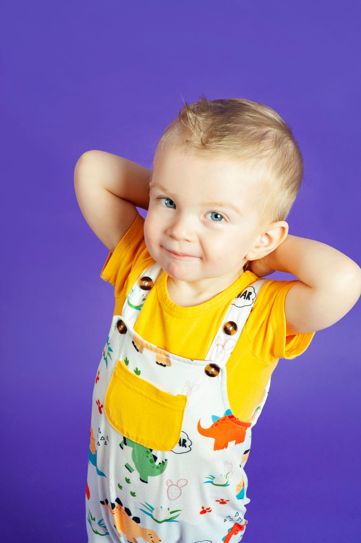 Cute toddler boy in dinosaur overalls and yellow shirt posing against purple background.
