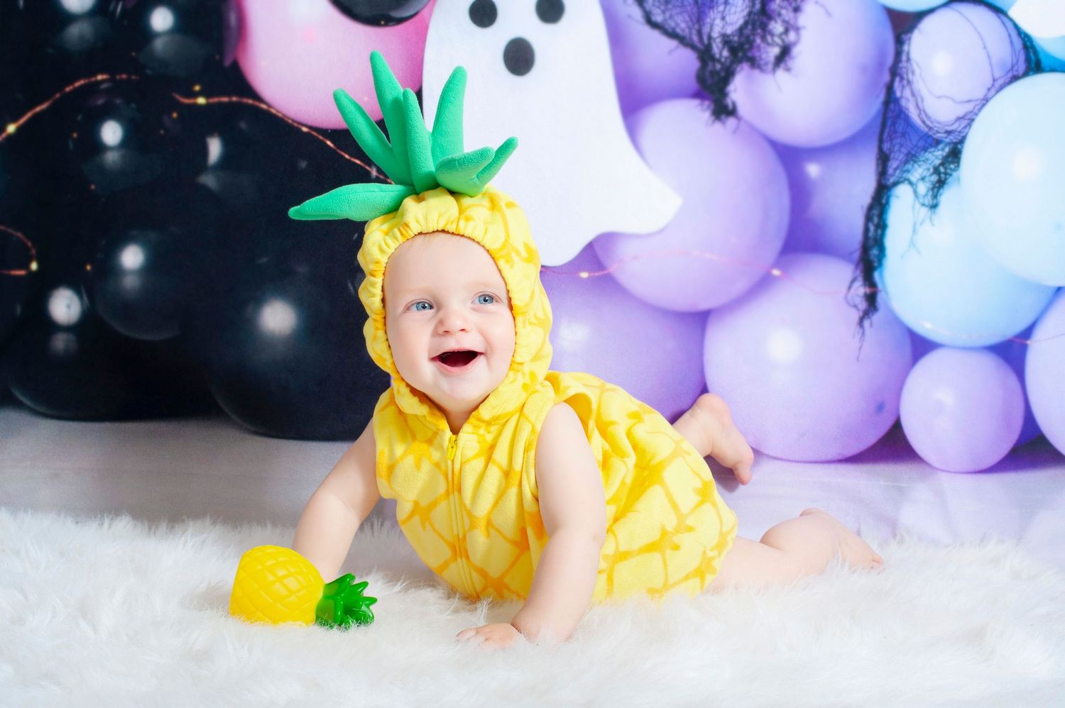 A laughing baby in a yellow pineapple Halloween costume sits on a white rug surrounded by colorful balloons.