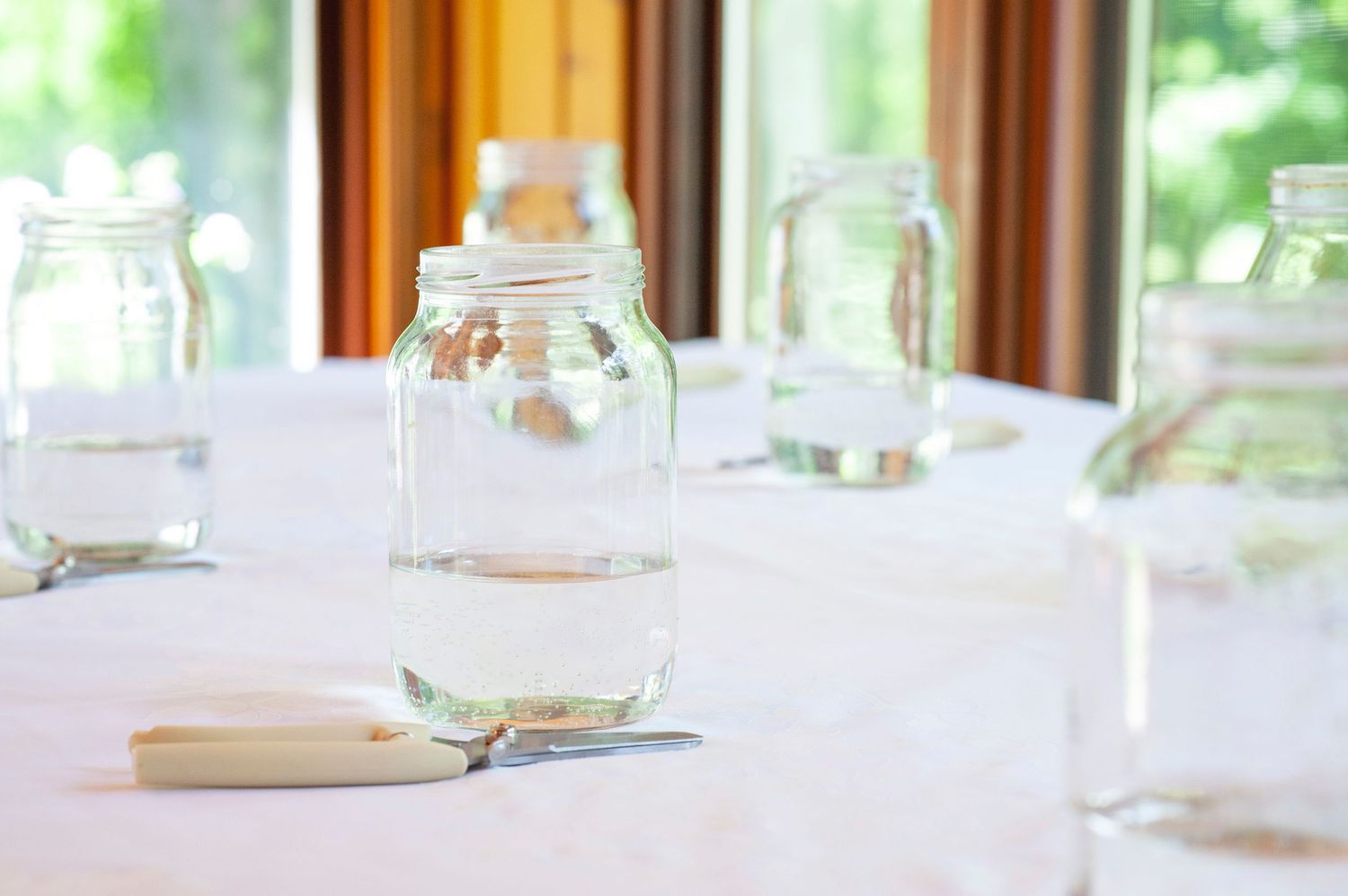 Clear glass mason jars on white tablecloth near sunlit window for rustic table setting.