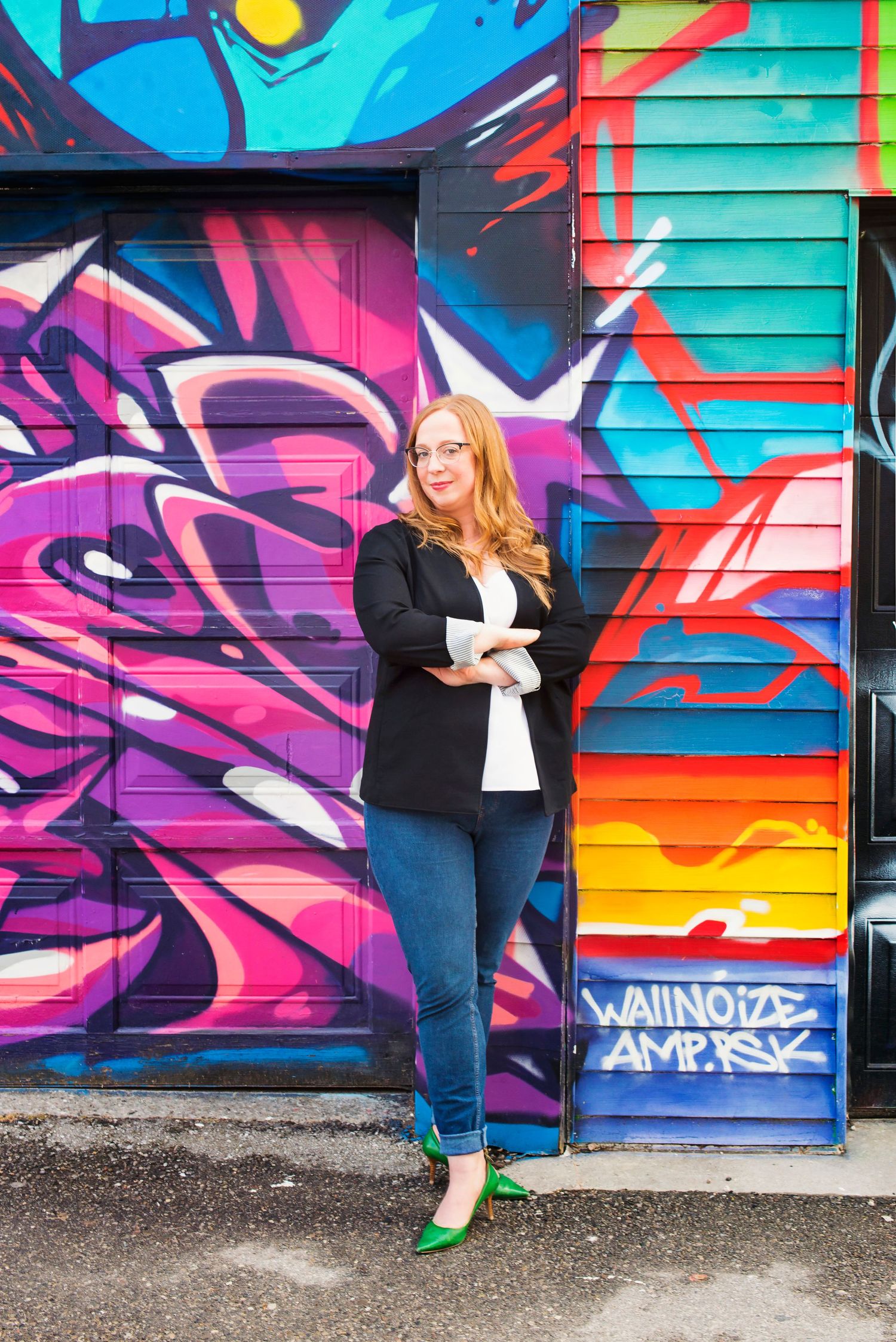 Confident redhead woman in black blazer and green heels poses before a colorful graffiti wall.