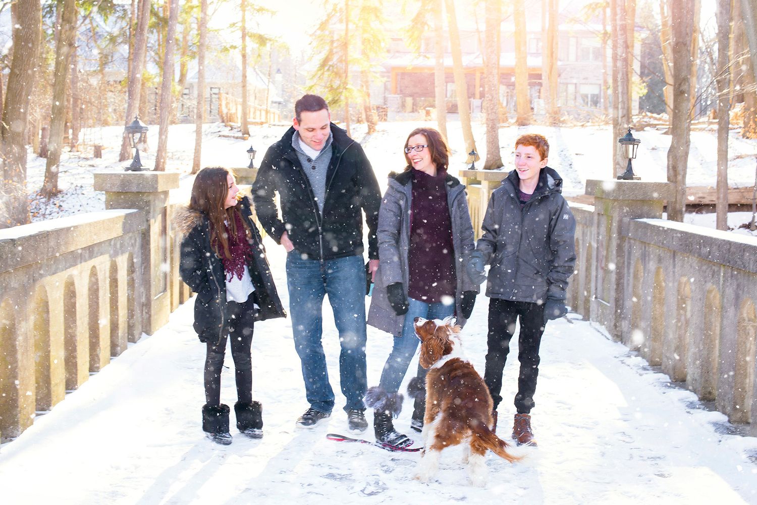 Happy family with dog walking on snowy bridge in winter park with falling snow.