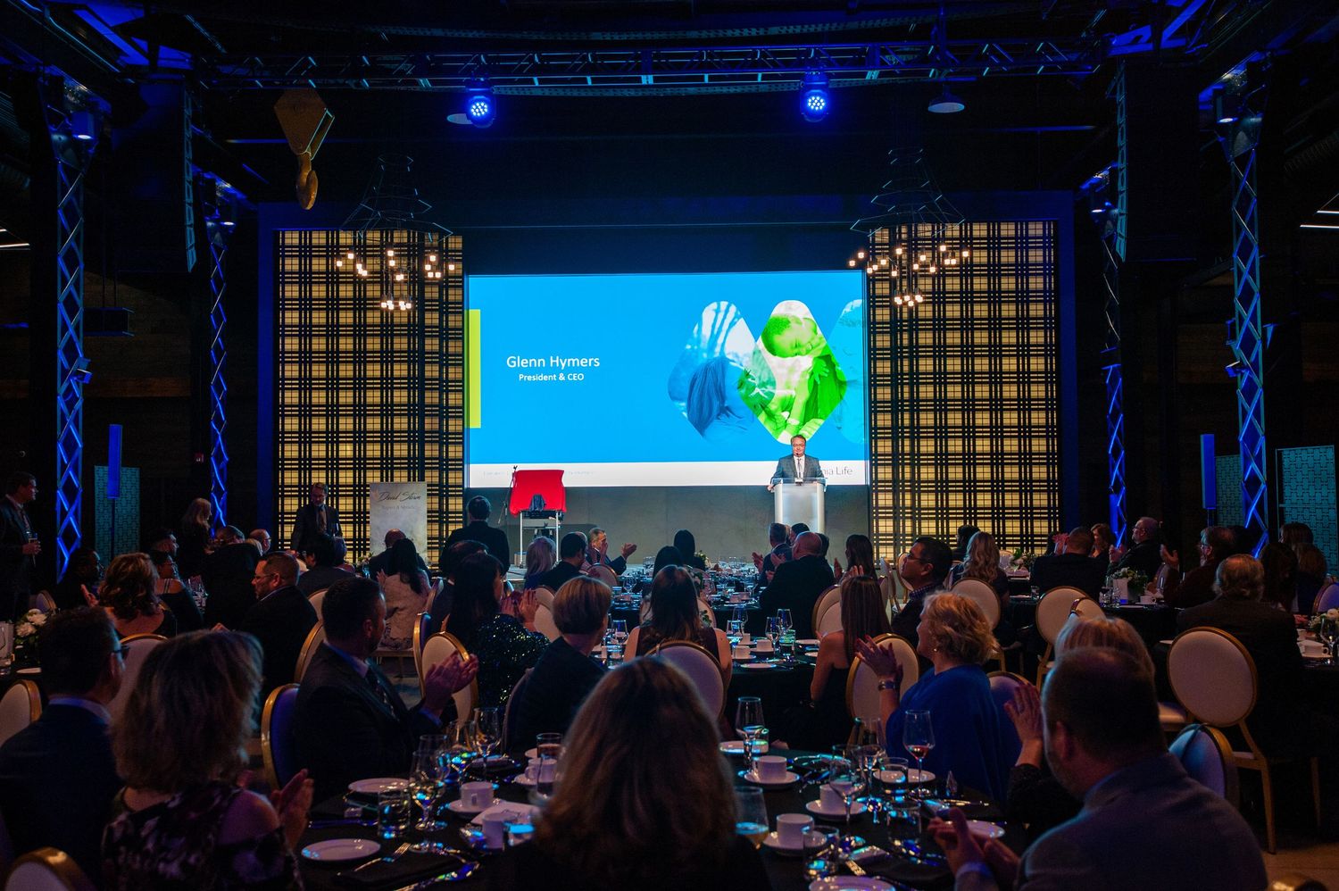 Audience seated at gala dinner tables facing a large presentation screen on stage.