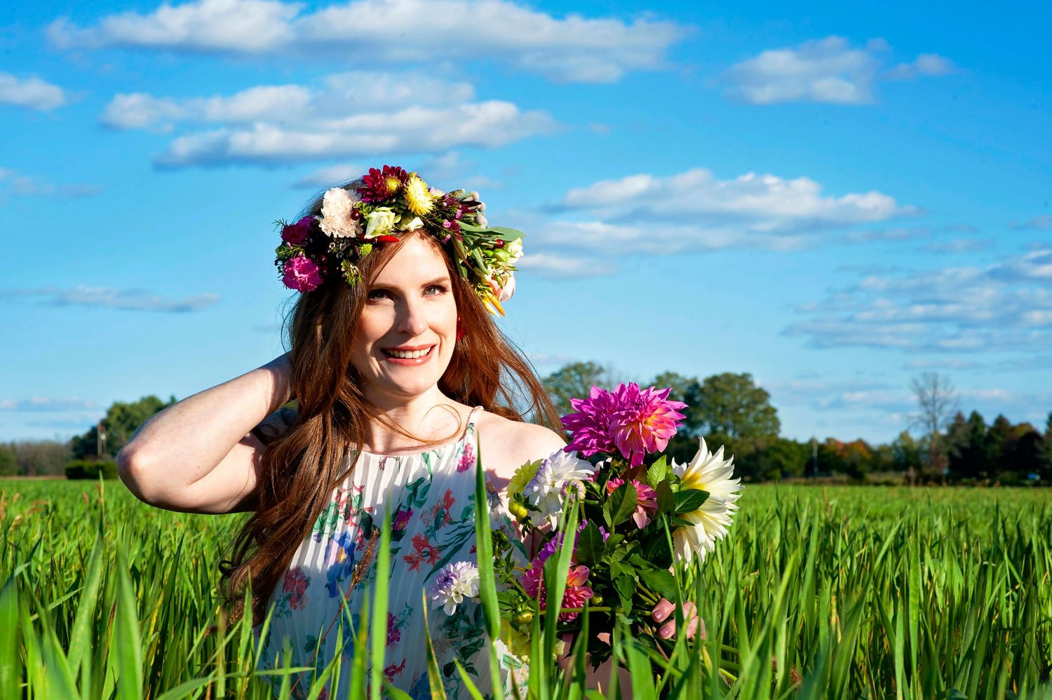 Woman with floral crown holding flowers in a green field under blue sky.