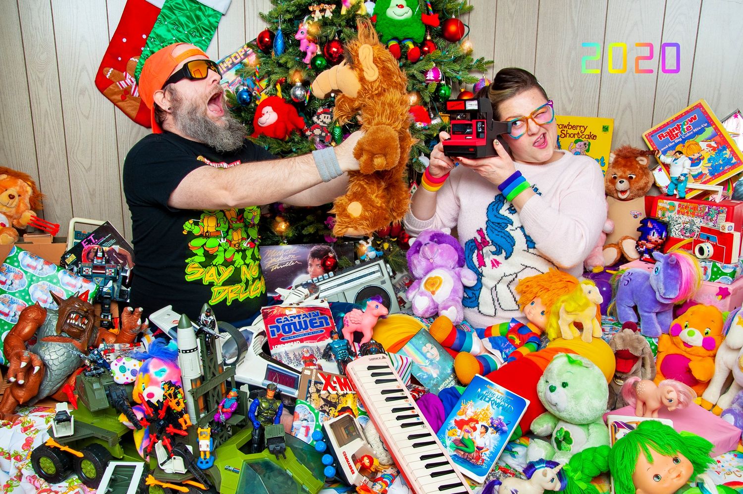 Two adults surrounded by colorful toys and stuffed animals near a Christmas tree celebrating 2020.