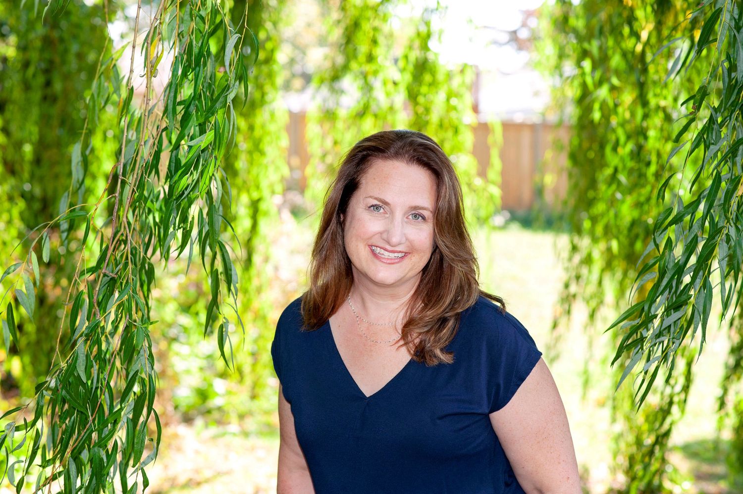 Brunette woman in navy top smiling under weeping willow tree branches outdoors.