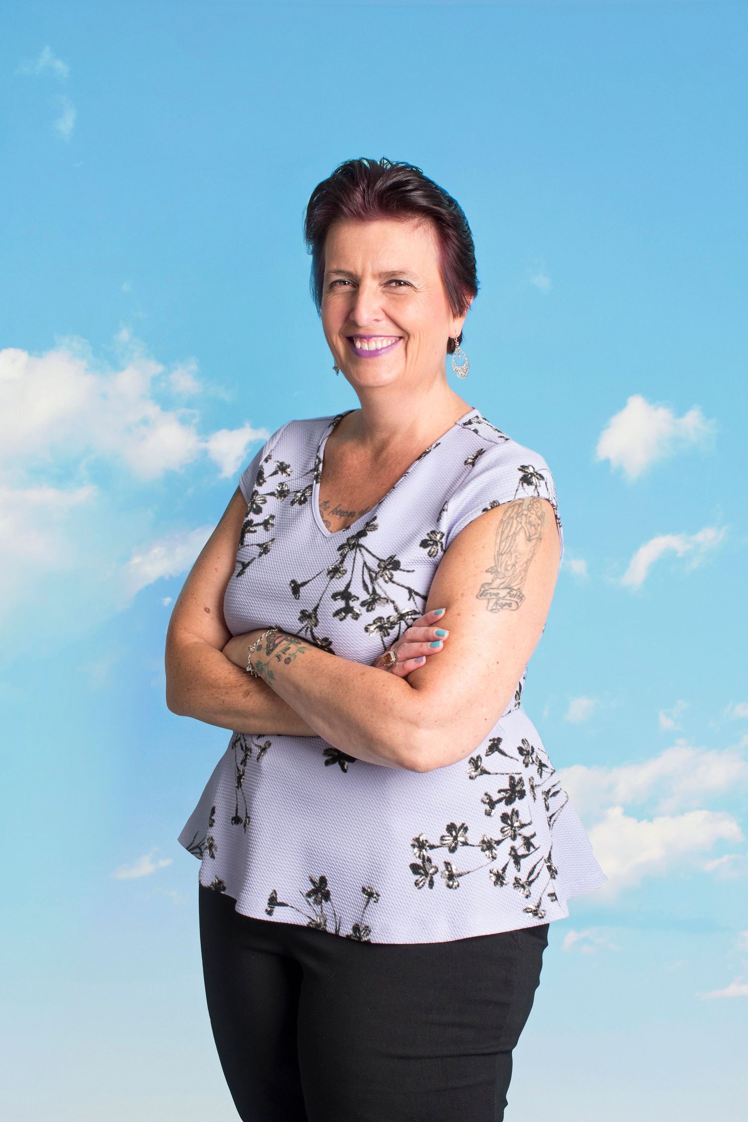 Smiling middle-aged woman with tattoo posing arms crossed against blue sky background.
