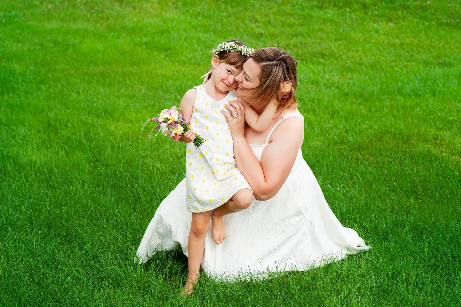 Mother kneeling on green grass hugging young daughter holding small bouquet outdoors.