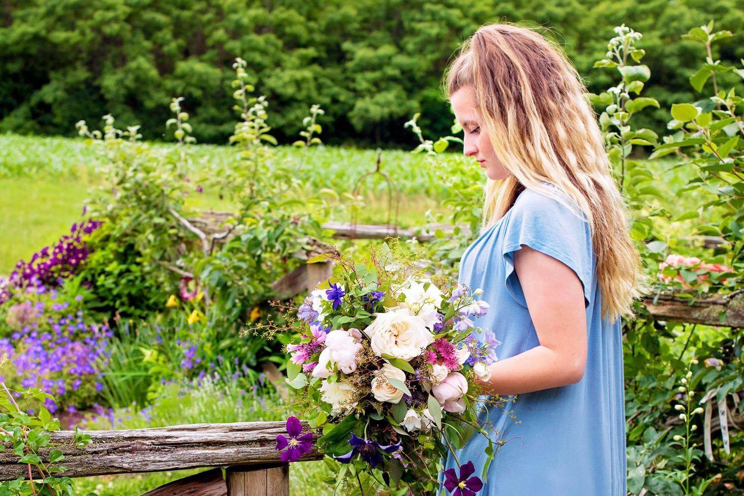 Blonde woman in blue dress admiring a garden bouquet of roses in a lush green garden.
