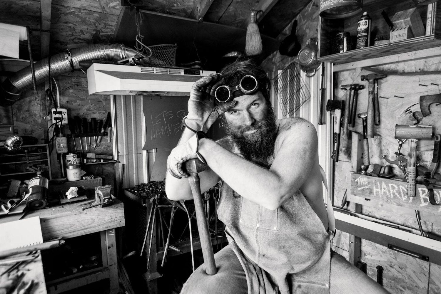 Shirtless bearded man with steampunk goggles working in a rustic tool-filled workshop, black and white.
