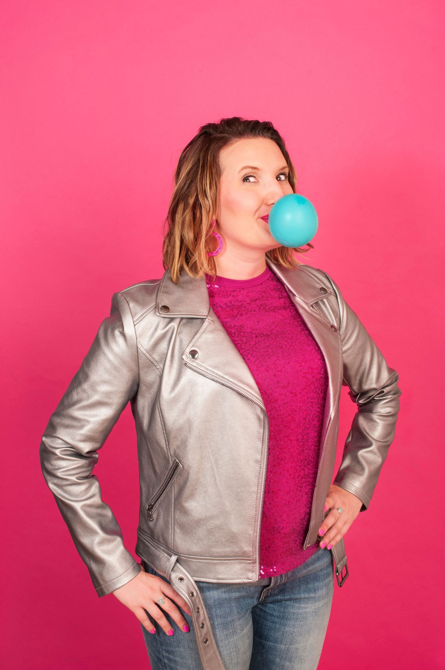 Woman in silver leather jacket blowing a blue bubble gum bubble against a bright pink background.