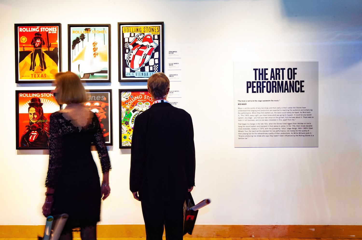 Couple views Rolling Stones concert posters beside The Art of Performance exhibit sign.