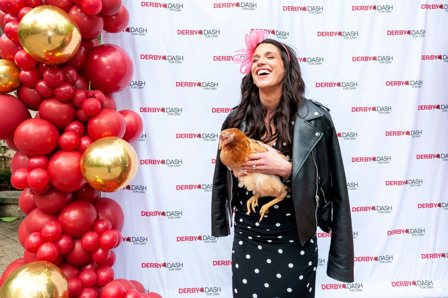 Woman in leather jacket holding a small dog laughing at a Derby Cash branded step-and-repeat backdrop.