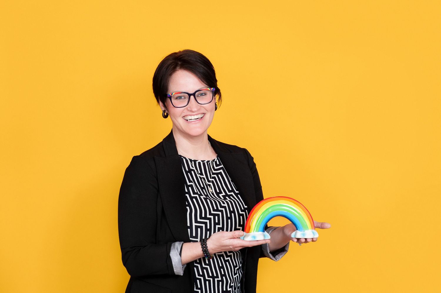 Smiling woman in black blazer holding a colorful rainbow decoration on yellow background.