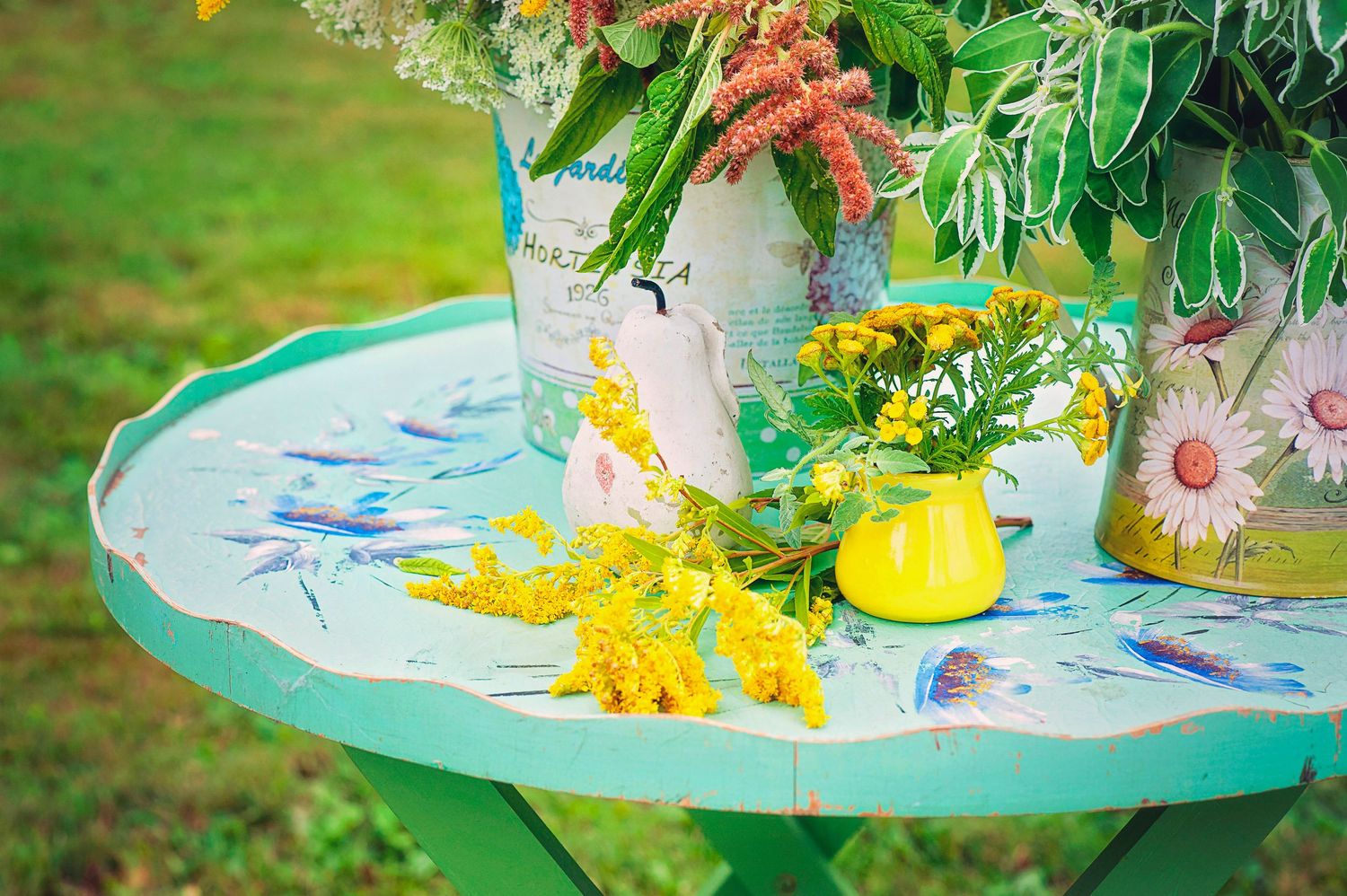 Green vintage table with yellow flowers and floral vases arranged in a garden setting.
