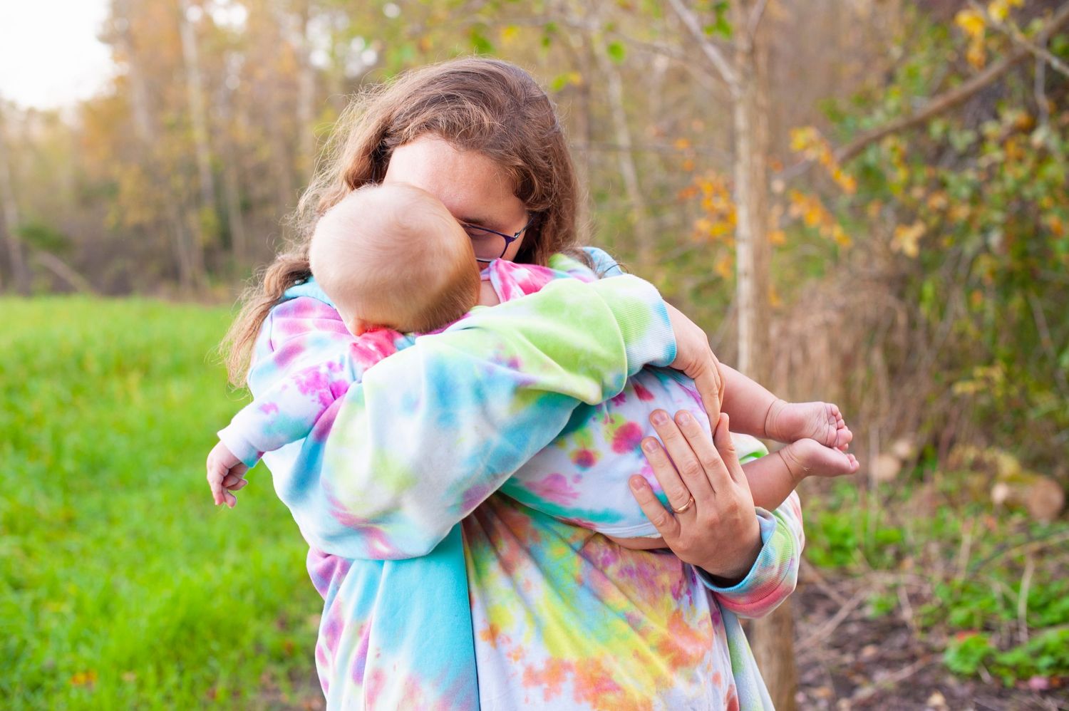 Person in tie-dye hoodie tenderly hugging a baby outdoors in nature.