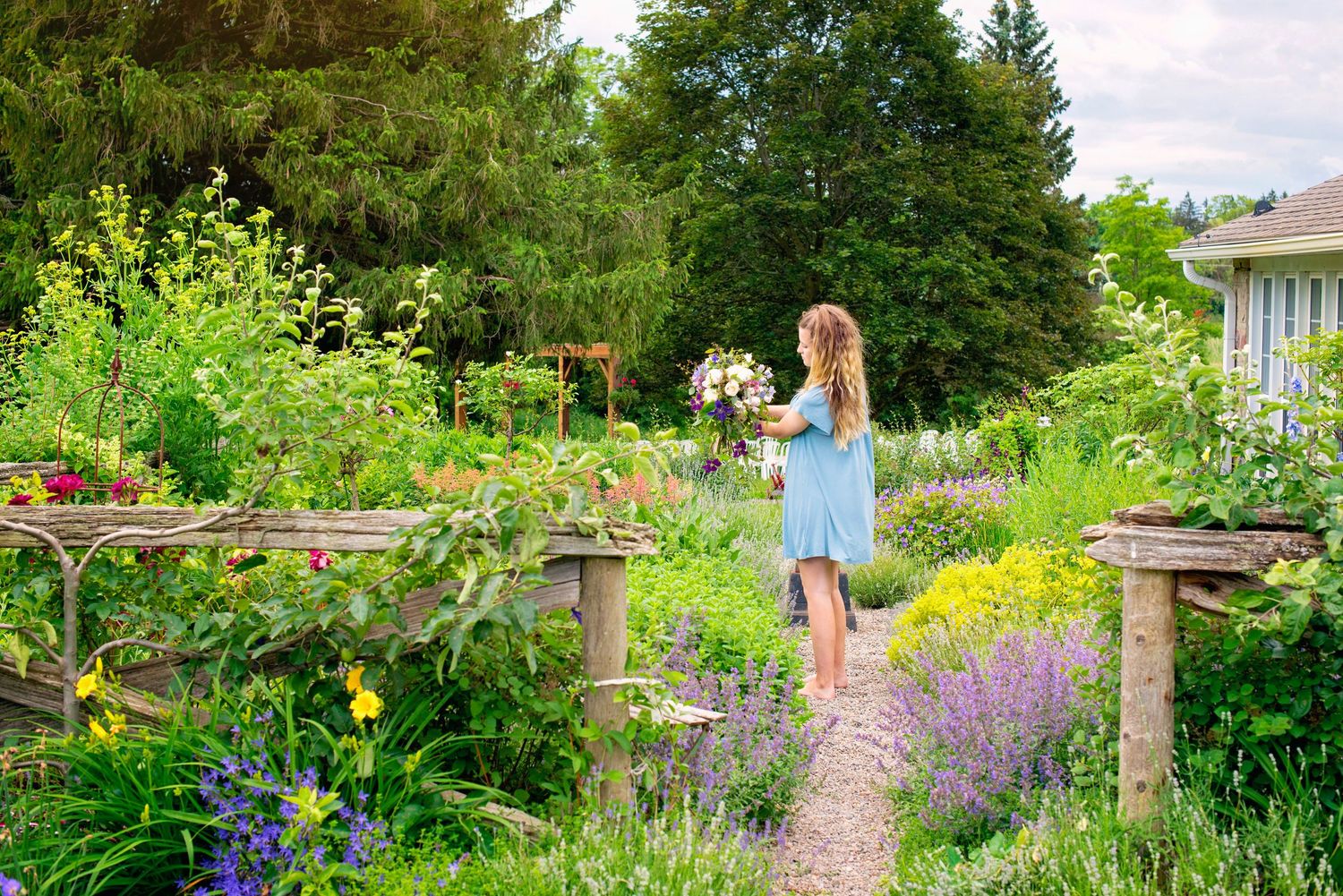 Young girl in blue dress walking through a lush cottage garden path holding flowers.