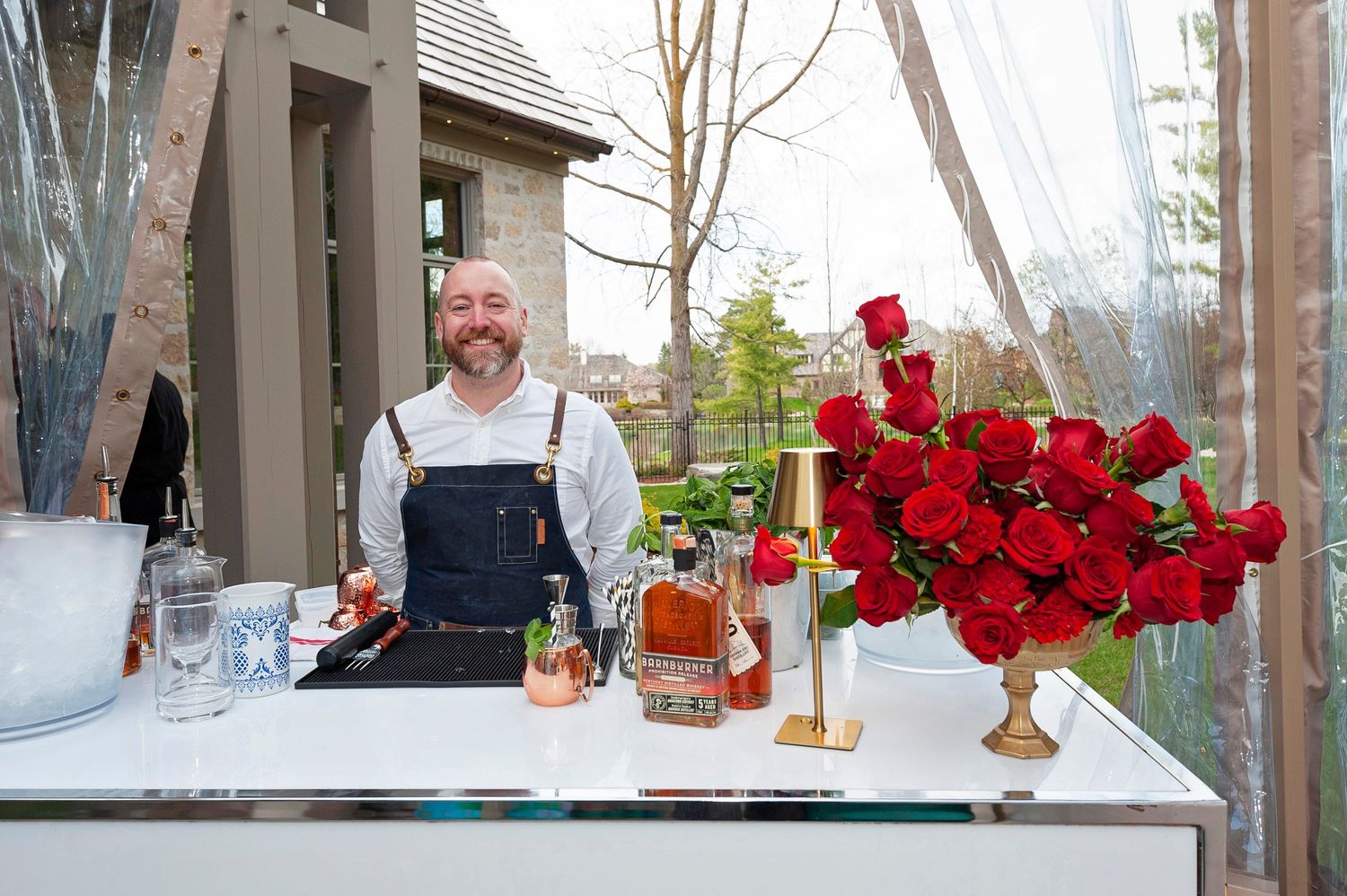 Smiling bartender standing behind a white bar with red roses and whiskey bottles at an upscale party event.