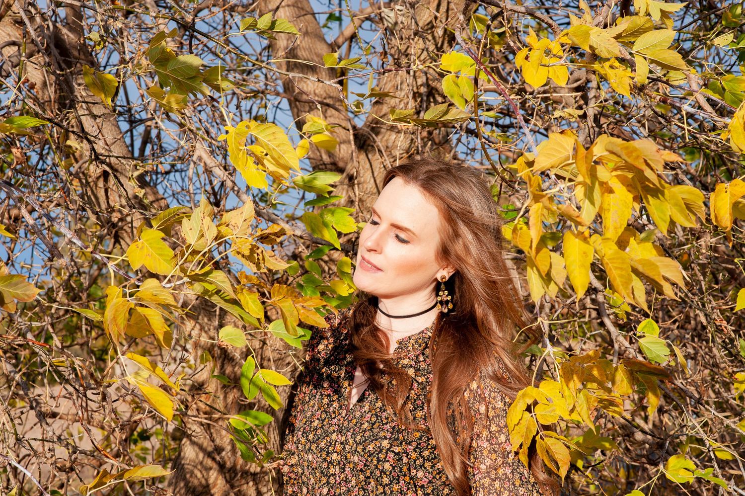 Young woman with long brown hair standing among golden autumn leaves in bright sunlight.