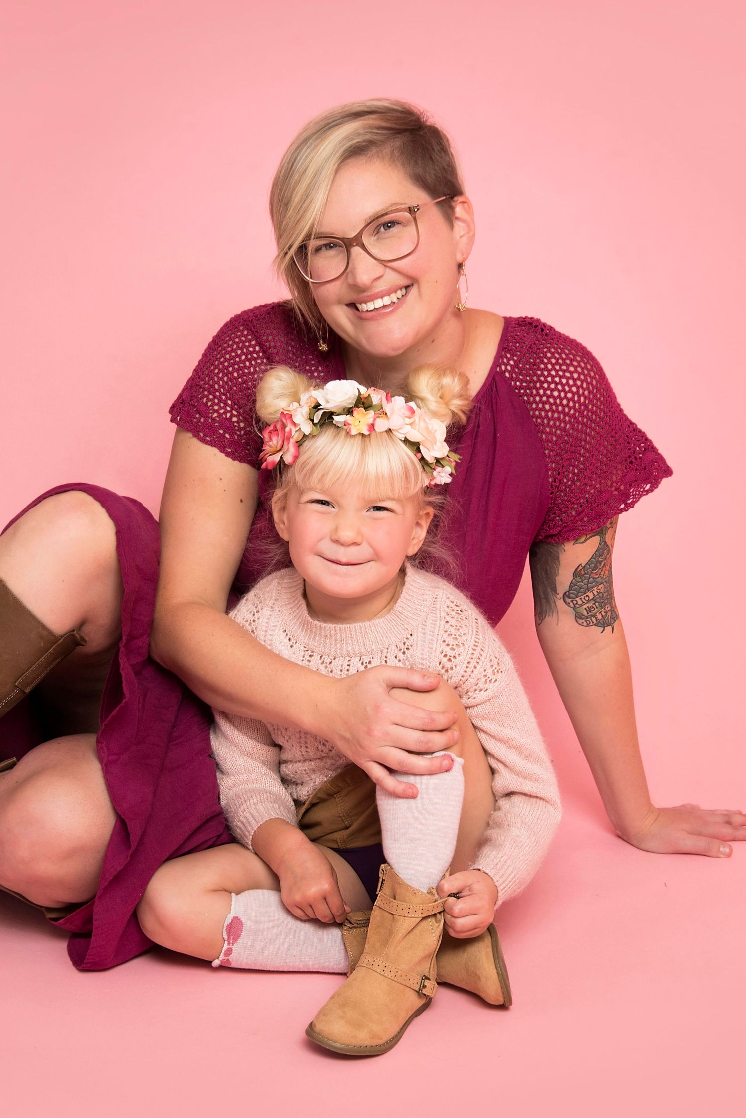 Smiling mother with glasses hugging young daughter wearing floral crown on pink studio background.