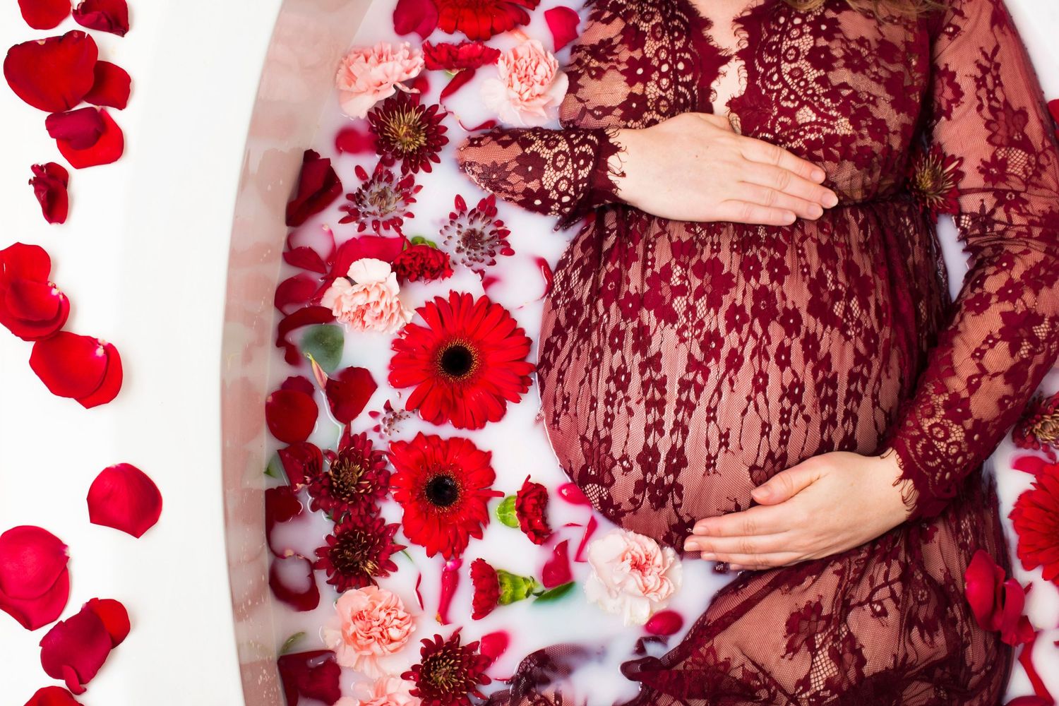 Pregnant woman in red lace dress lying in floral milk bath with red poppies and rose petals.