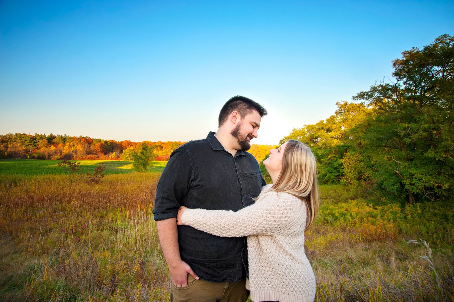 Loving couple embracing in an open field during golden hour with colorful fall foliage in the background.