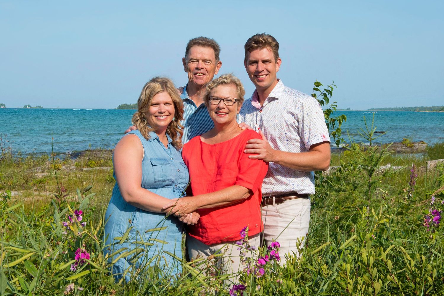 Family of four smiling outdoors near a scenic waterfront with lush greenery and blue ocean in the background.