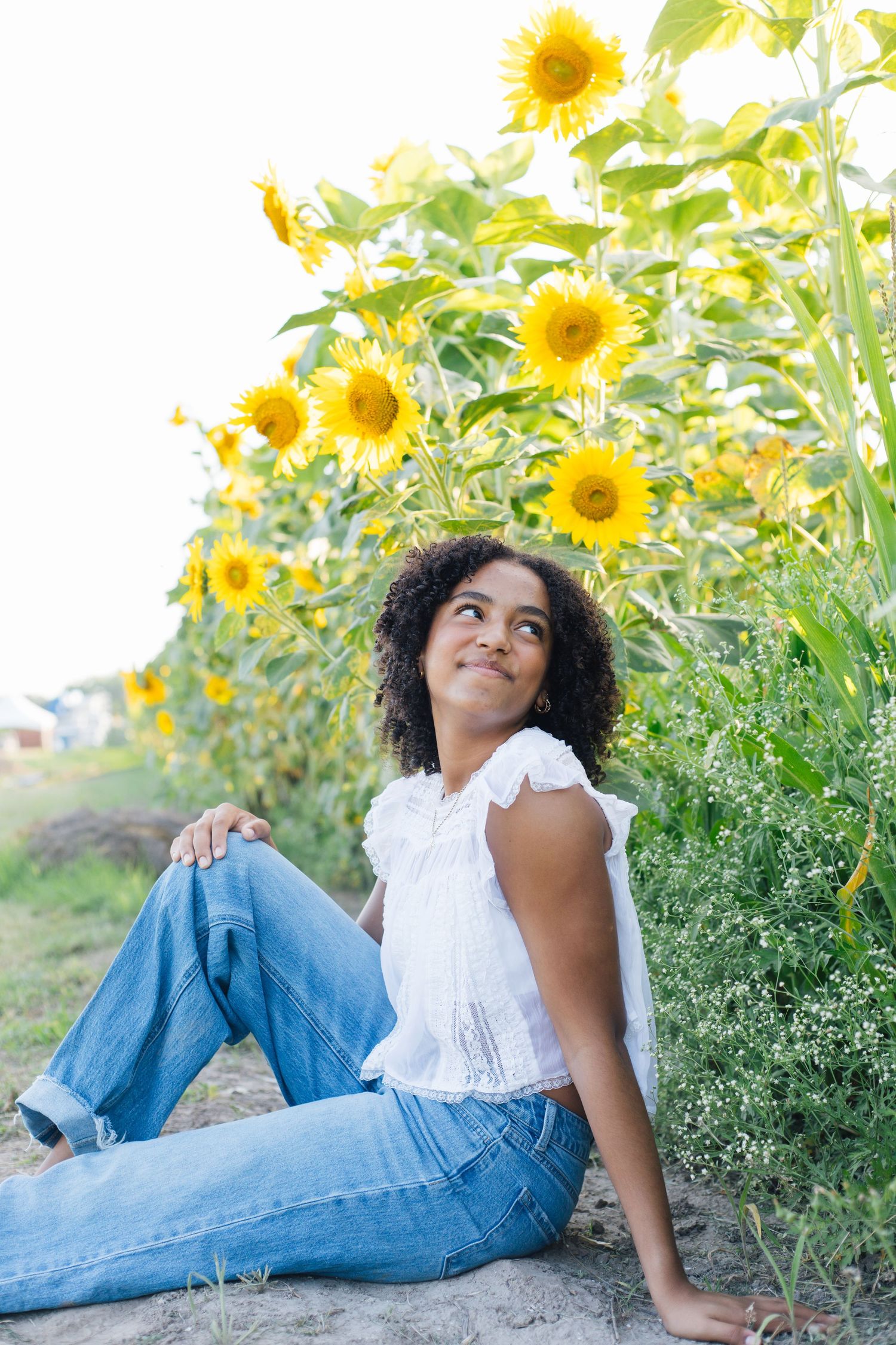 Young woman in white top and jeans sitting by sunflower field, looking up thoughtfully on a sunny day.