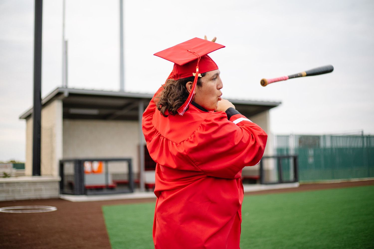 Graduate in red cap and gown swings a baseball bat on a field, celebrating graduation day.