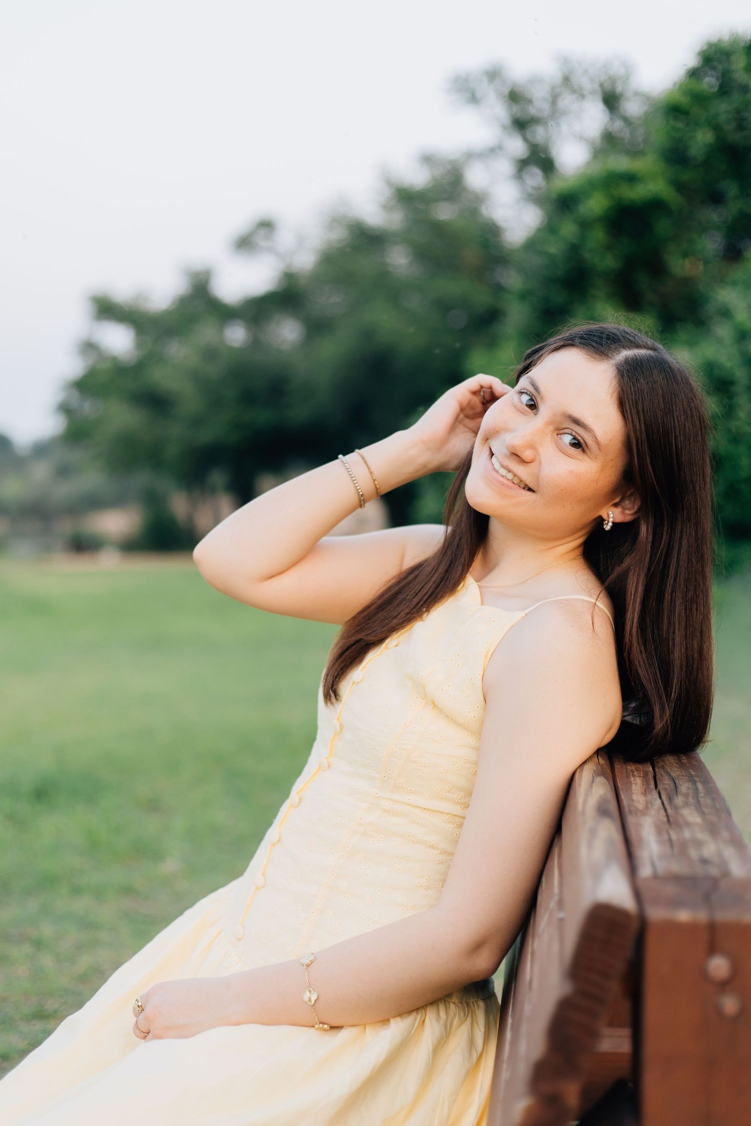 Young woman in yellow dress sitting on wooden bench in park, smiling with hand in hair. Senior Portraits Round Rock TX