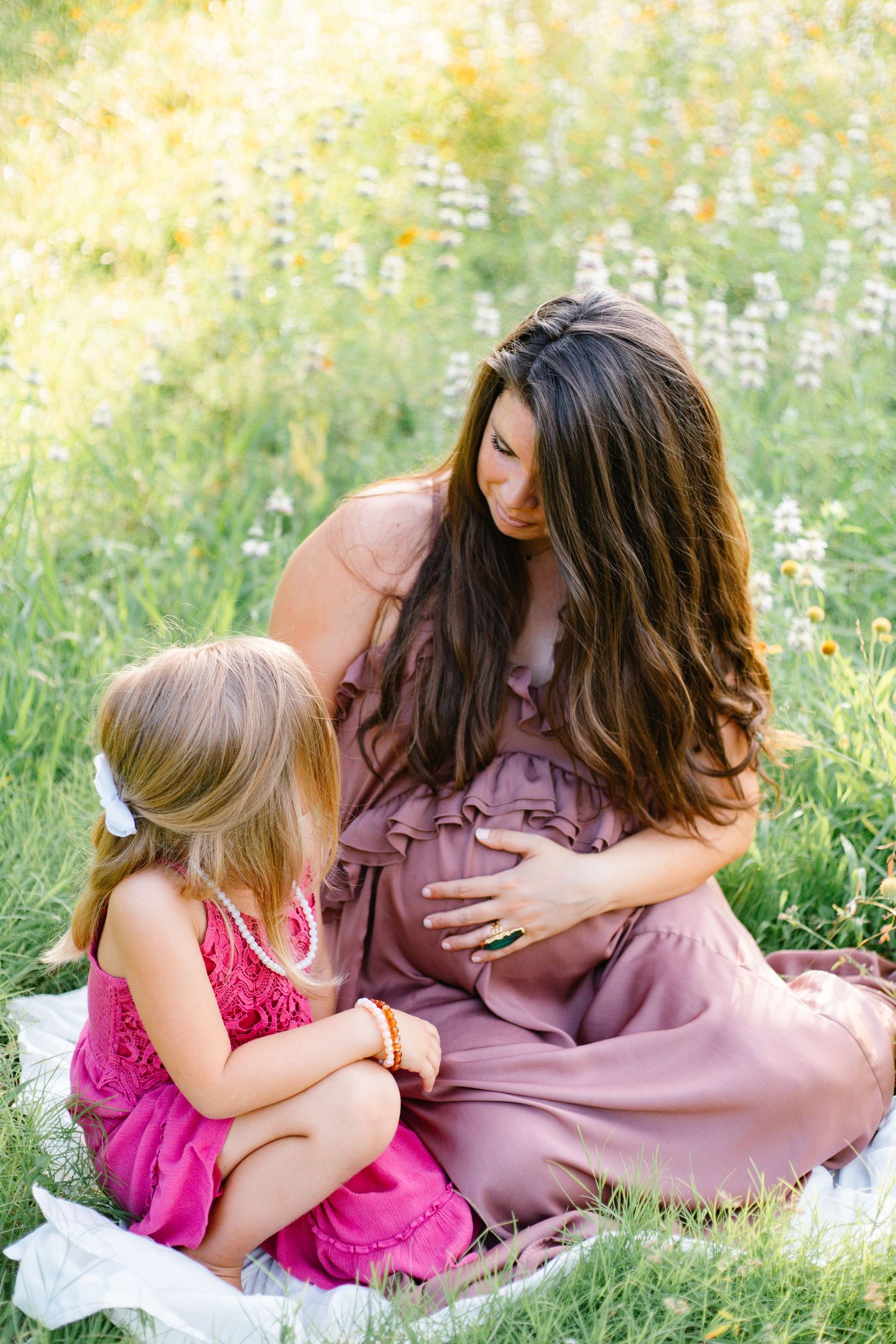 Pregnant mother in mauve dress sits in sunny wildflower field with young blonde daughter touching bump.
