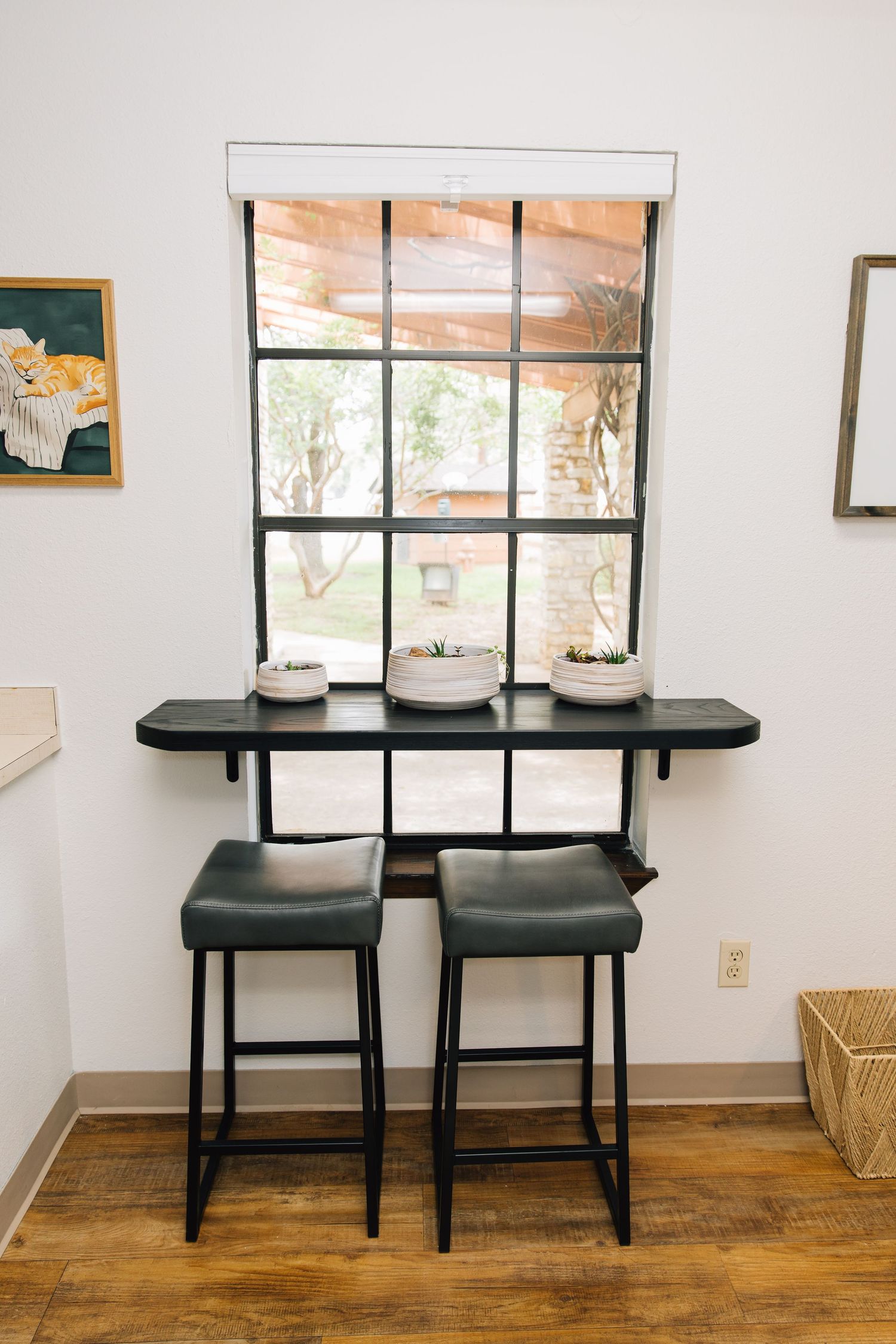 Two black bar stools sit beneath a wall-mounted narrow desk below a multi-pane window with small potted plants on top.