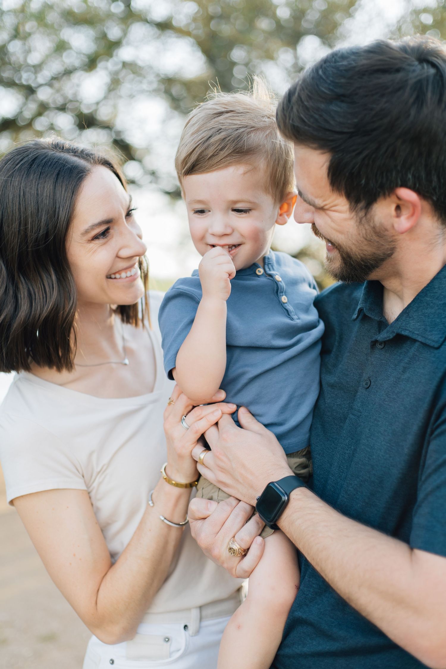 Happy young parents holding their laughing toddler son outdoors in a sunlit park setting.