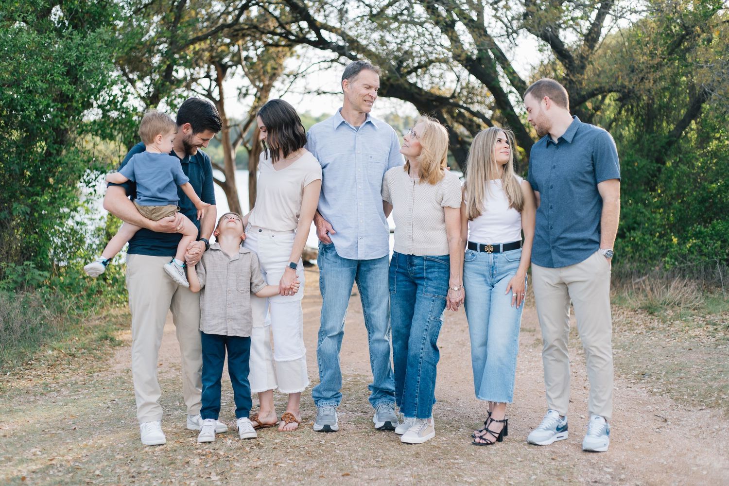Large family group portrait outdoors on a dirt path surrounded by green trees, wearing coordinated neutral outfits.