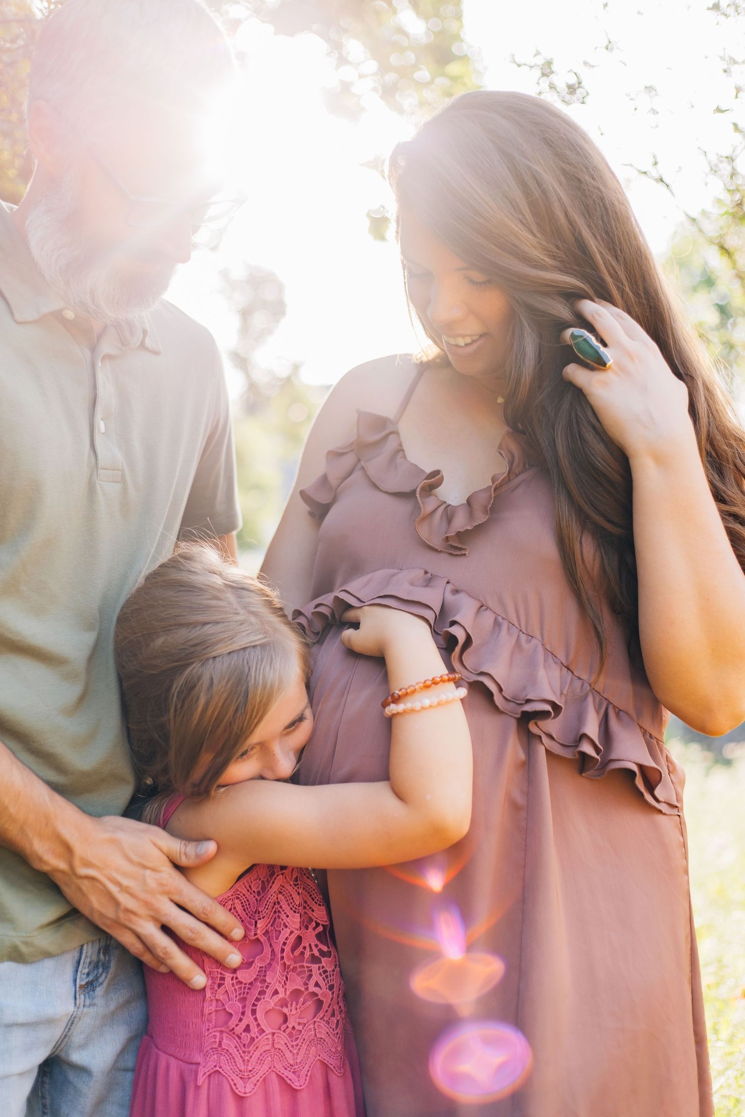 Pregnant woman in mauve dress smiles at daughter hugging her bump beside father in golden sunlight.
