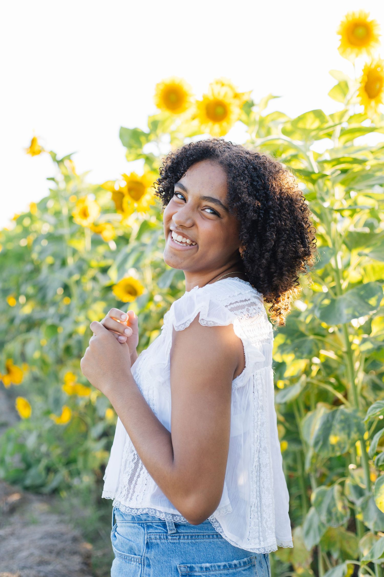 Smiling young woman with curly hair in white top and jeans standing in a vibrant sunflower field on a sunny day.