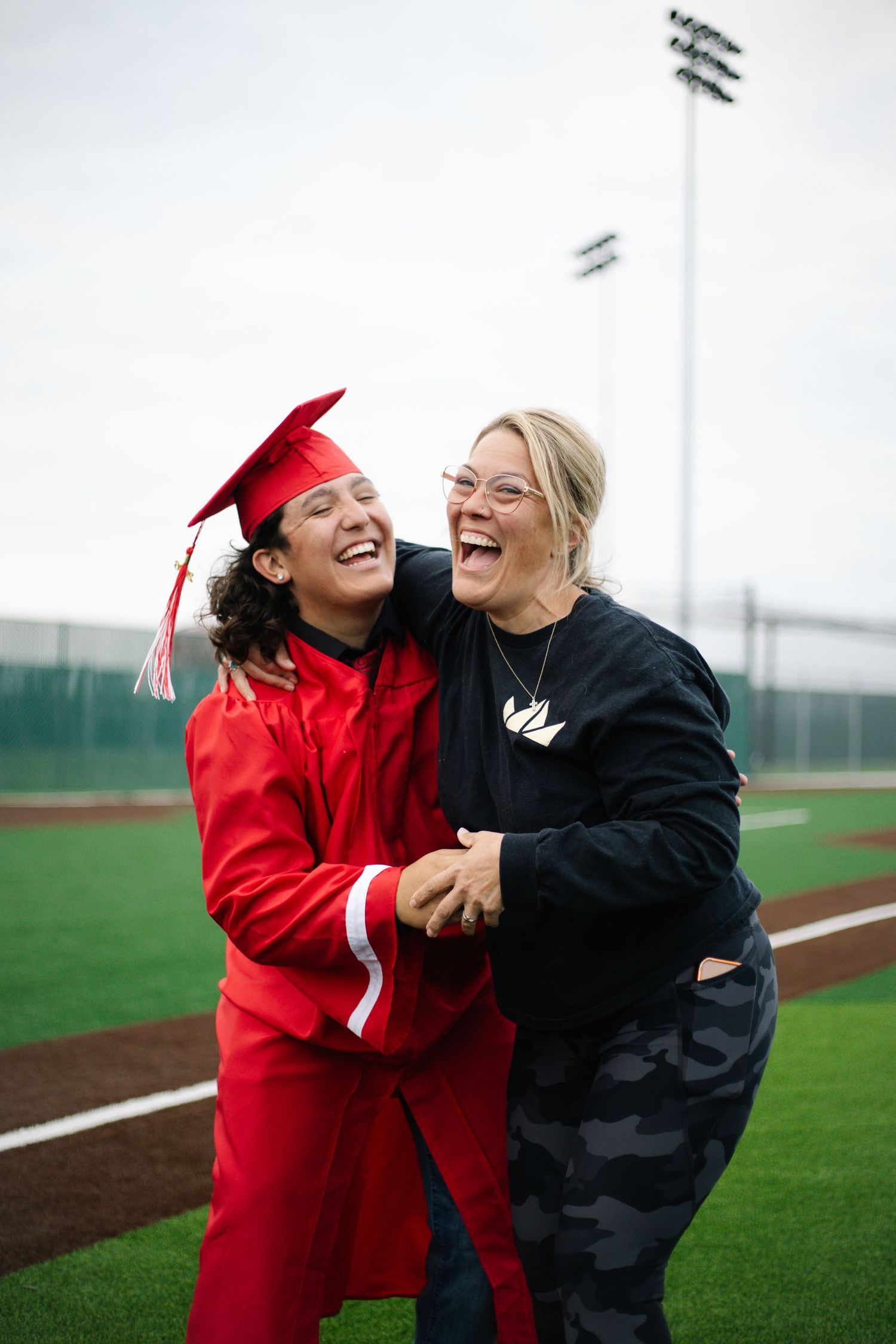 Two women laughing and embracing on a sports field, one wearing a red graduation cap and gown.