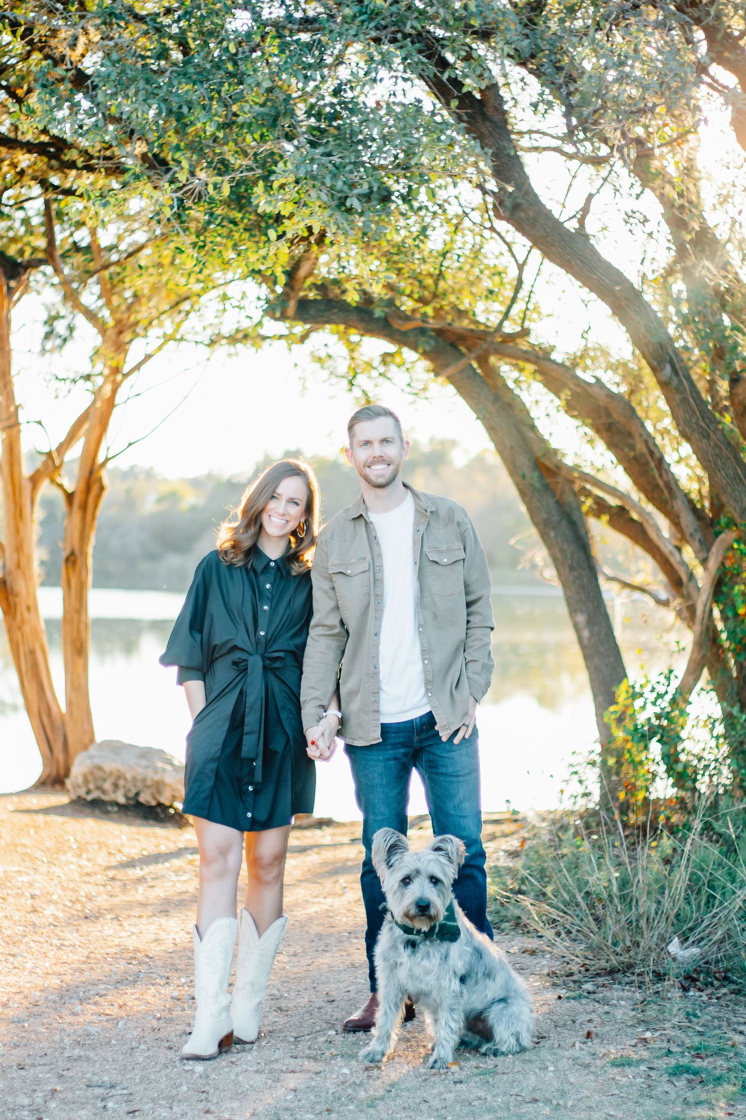 A couple poses with their fluffy grey dog under trees near a lake during a sunset photo session.